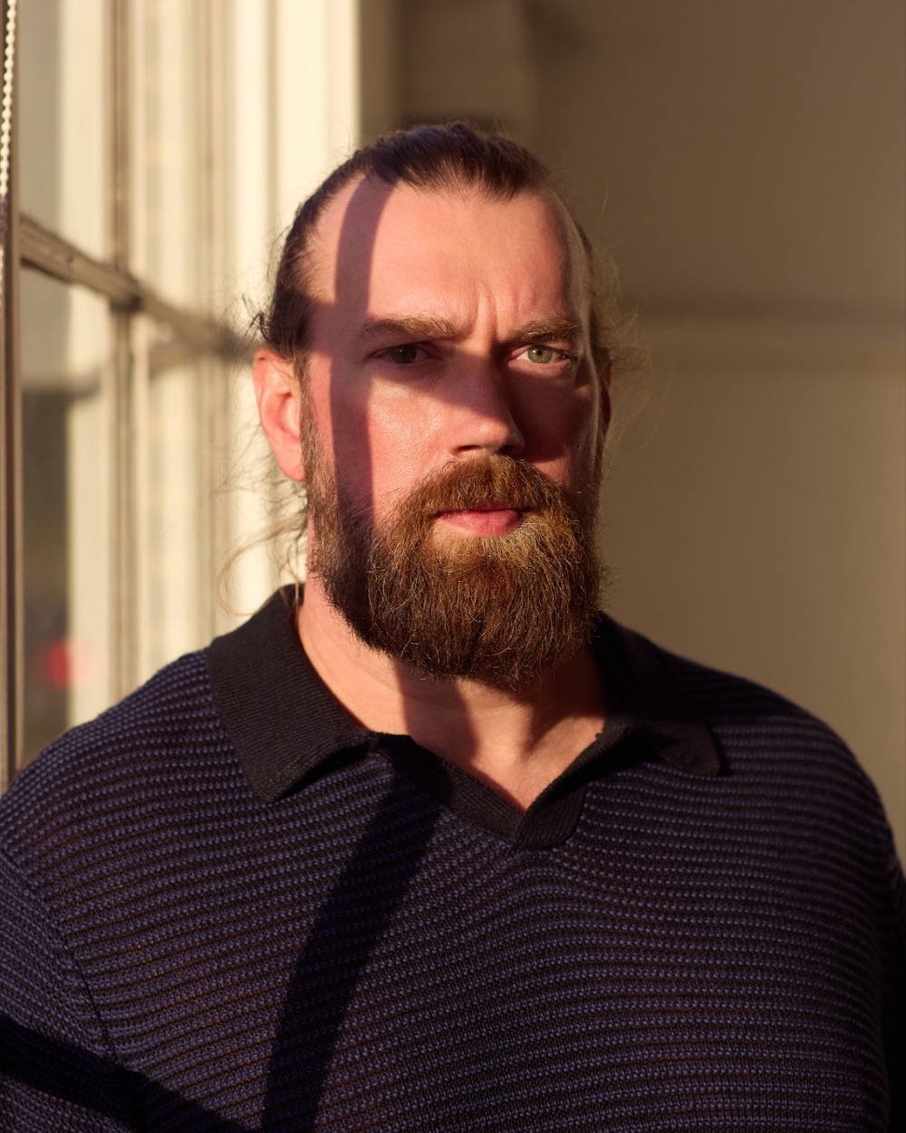 Brandon Wilburn by a window in late light, the window frame casting shadows across his face